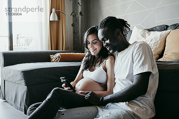 Junger Mann mit schwangerer Frau  die ein Smartphone benutzt  während sie zu Hause im Wohnzimmer sitzen