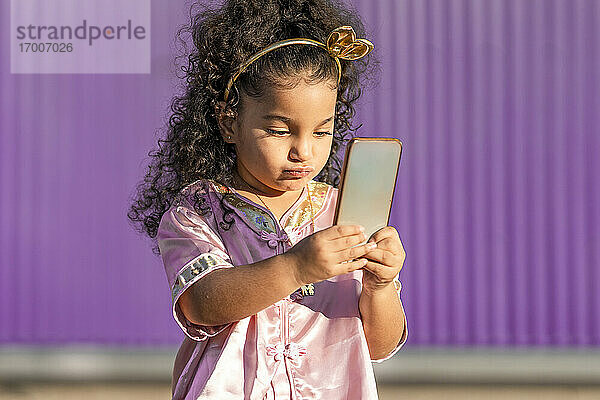 Nettes Mädchen  das sein Handy benutzt  während es gegen eine lila Wand steht