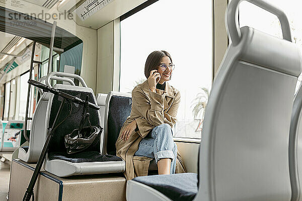 Lächelnde Frau  die mit einem Mobiltelefon spricht  während sie mit einem elektrischen Roller in der Straßenbahn sitzt