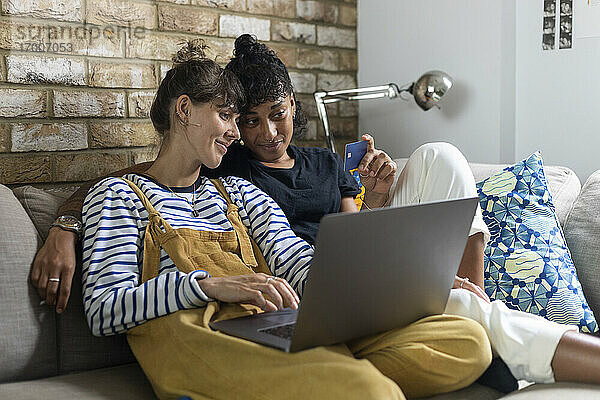 Frau mit Laptop und Kreditkarte in der Hand  während sie bei ihrer Freundin zu Hause sitzt