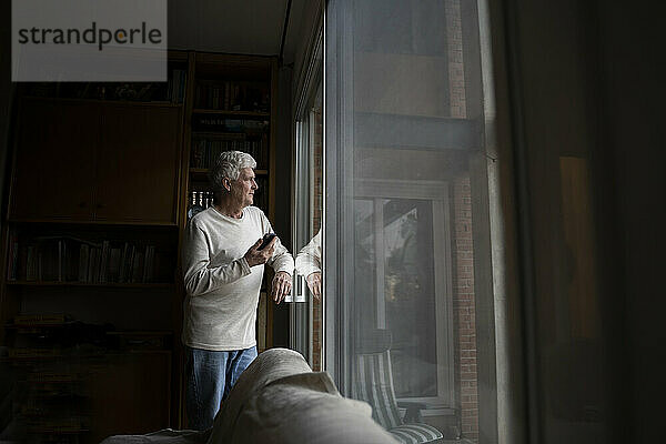 Älterer Mann mit Mobiltelefon  der wegschaut  während er zu Hause am Fenster steht