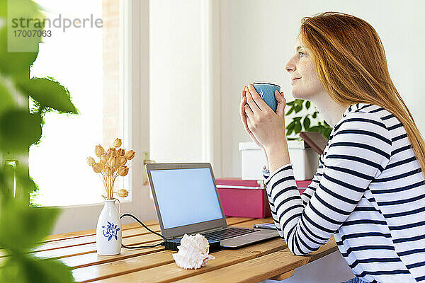 Junge rothaarige Frau mit Laptop trinkt Kaffee  während sie zu Hause sitzt