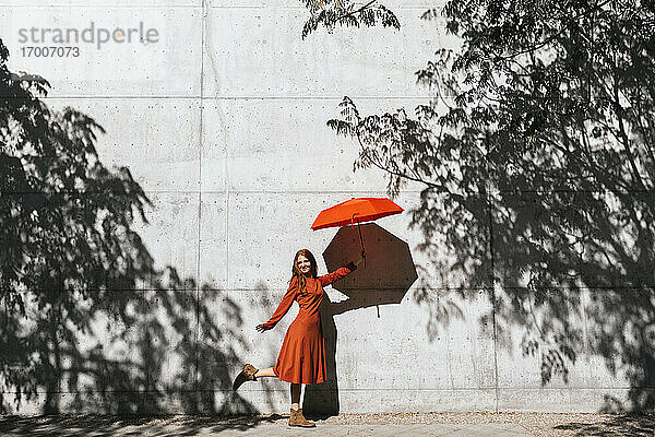 Lächelnde Frau  die einen roten Regenschirm hält  während sie vor einer Baumschattenwand steht