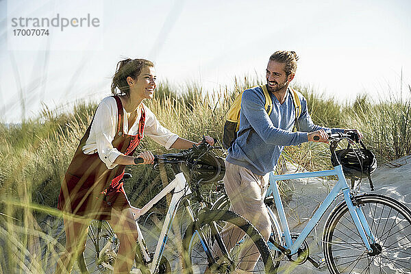 Lächelnde Freundin und Freund gehen mit Fahrrädern am Strand spazieren