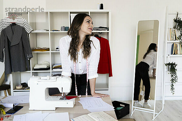 Weibliche Modedesignerin schaut weg  während sie sich im Atelier auf den Schreibtisch stützt