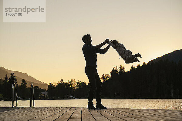 Silhouetten von Vater und kleiner Tochter  die in der Abenddämmerung am Ende eines Stegs am Seeufer spielen
