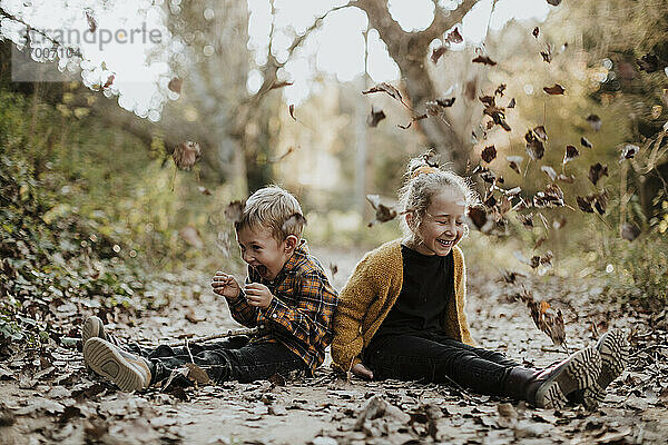Fröhliches Mädchen und Junge spielen mit trockenen  gefallenen Blättern  während sie auf einem Fußweg im Wald sitzen