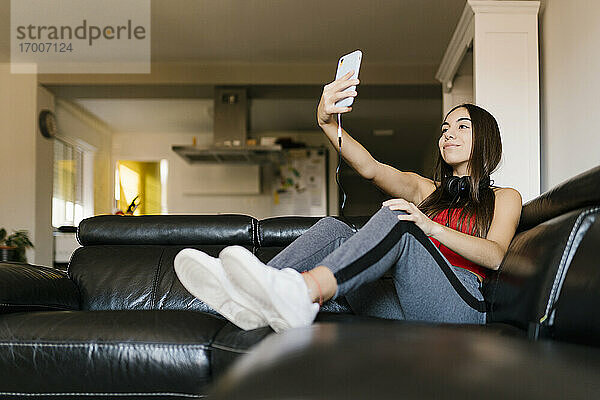 Lächelndes Teenager-Mädchen  das ein Selfie mit seinem Mobiltelefon macht  während es zu Hause auf dem Sofa sitzt
