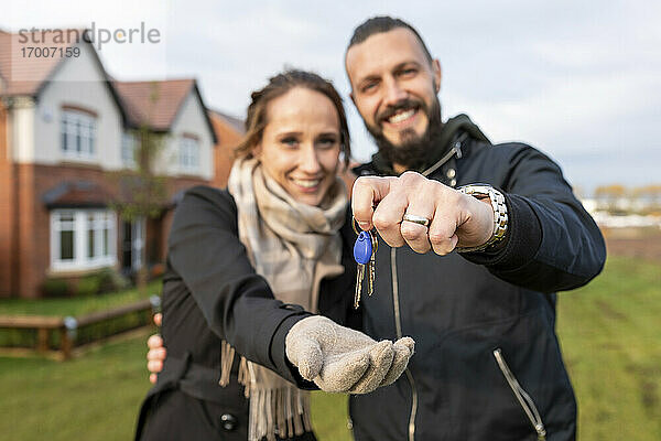 Glücklicher Mann  der seiner Freundin die Schlüssel für sein neues Haus gibt  während er im Vorgarten steht