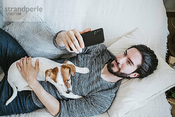 Junger Mann  der ein Selfie mit seinem Handy macht  während er mit seinem Hund zu Hause auf dem Sofa liegt