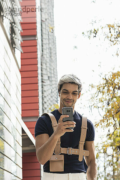 Lächelnder Mann mit Hand in der Tasche  der sein Smartphone in der Stadt benutzt
