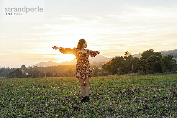 Junge Frau im Sonnenkleid mit ausgestreckten Armen gegen den Himmel bei Sonnenuntergang