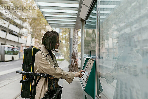 Frau mit Instrumentenkoffer  die einen Fahrkartenautomaten benutzt  während sie mit einem Elektroroller an einer Straßenbahnhaltestelle in der Stadt steht