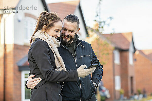Lächelnder Mann  der seine Freundin mit dem Smartphone ansieht  während er vor neuen Häusern steht