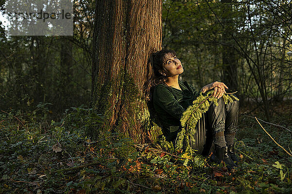 Schöne Frau mit Zweig träumt Tag  während lehnt sich auf Baumstamm in öffentlichen Park