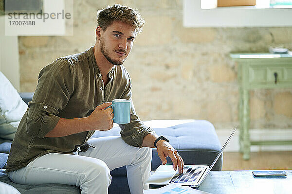 Männlicher Freiberufler hält Kaffeetasse  während er zu Hause am Laptop sitzt