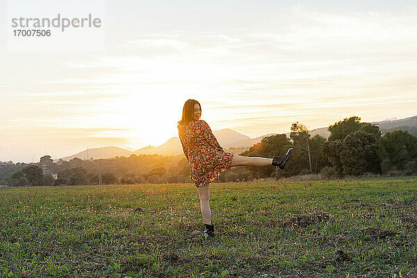Fröhliche junge Frau auf einem Bein stehend auf einer Wiese bei Sonnenuntergang