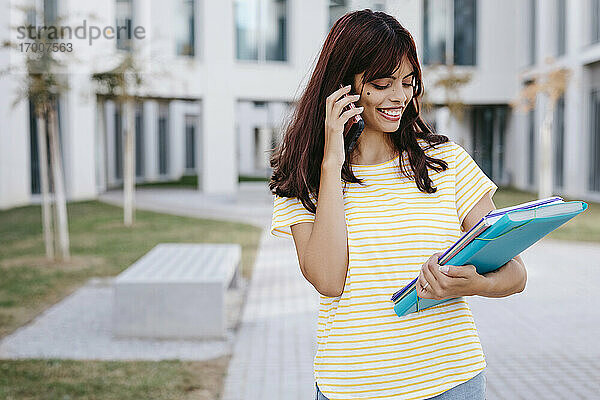 Lächelnde lateinamerikanische Studentin  die mit ihrem Handy telefoniert  während sie sich Bücher und Akten ansieht