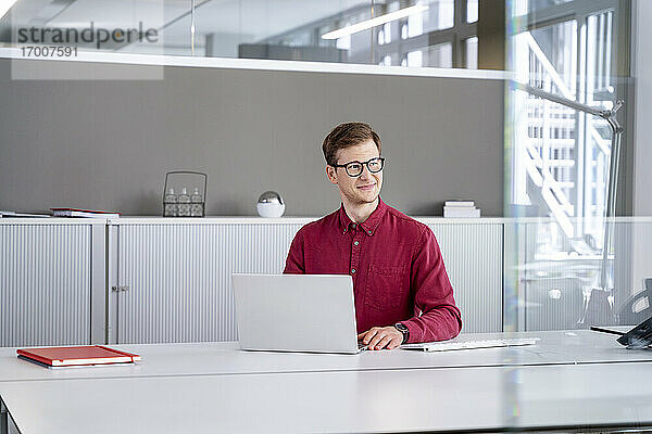 Lächelnder Geschäftsmann  der mit seinem Laptop im Büro sitzt und wegschaut