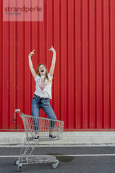 Mädchen schaukelt in einem Einkaufswagen vor einer roten Wand