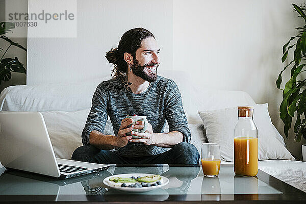 Lächelnder Mann mit Kaffeetasse  der wegschaut  während er zu Hause auf dem Sofa sitzt und frühstückt