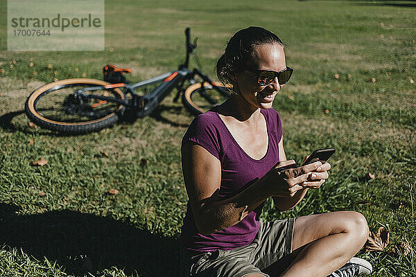 Lächelnde Frau  die ein Mobiltelefon benutzt  während sie auf einem elektrischen Mountainbike im Park sitzt