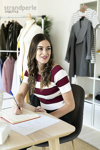 Lächelnde Modedesignerin  die mit einem Buch im Atelier sitzt und wegschaut
