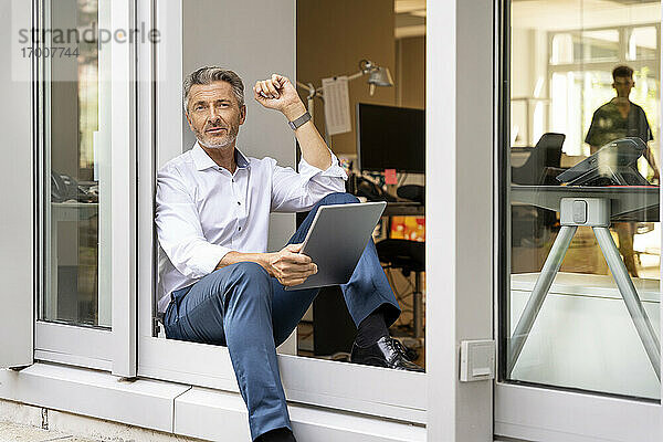Nachdenklicher Geschäftsmann mit digitalem Tablet  der wegschaut  während er an der Bürotür sitzt
