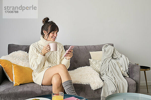 Lächelnde Frau  die ihr Smartphone benutzt  während sie zu Hause am Wintermorgen Kaffee trinkt