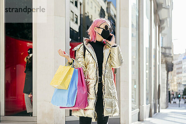 Frau mit Schutzmaske  die mit Einkaufstüten in der Stadt steht und mit dem Handy telefoniert