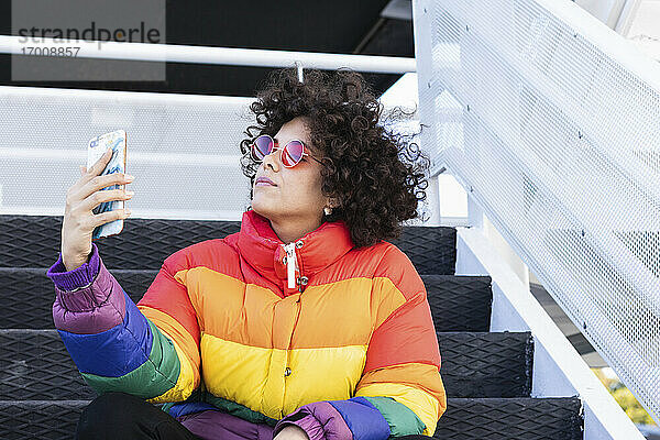 Frau mit bunter Jacke nimmt Selfie durch Handy beim Sitzen auf Stufen