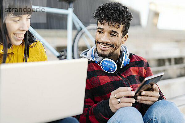 Lächelnde Freunde  die im Freien sitzend einen Laptop und ein digitales Tablet benutzen