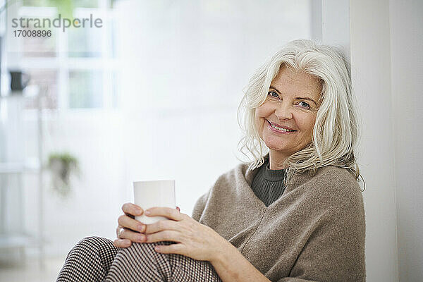 Glückliche ältere Frau hält eine Kaffeetasse  während sie zu Hause im Wohnzimmer sitzt