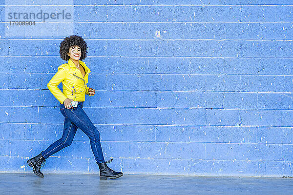 Glückliche Frau in gelber Lederjacke  die ihr Handy hält  während sie gegen eine blaue Wand läuft