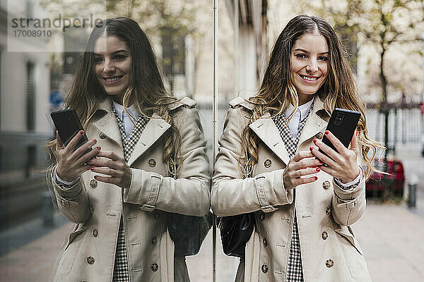 Glückliche Frau  die ein Mobiltelefon hält  während sie an einer Glaswand in der Stadt steht