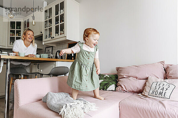 Kleines Mädchen springt auf dem Sofa  lächelnde Mutter im Hintergrund