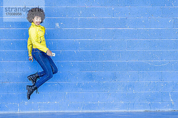 Glückliche modische Frau mit Smartphone springt gegen blaue Wand