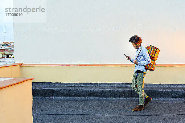 Junger Mann mit Rucksack und Smartphone beim Spaziergang auf der Terrasse