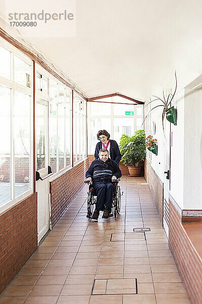 Weibliche Krankenschwester hilft behindertem Mann im Rollstuhl sitzend auf dem Flur eines Pflegeheims