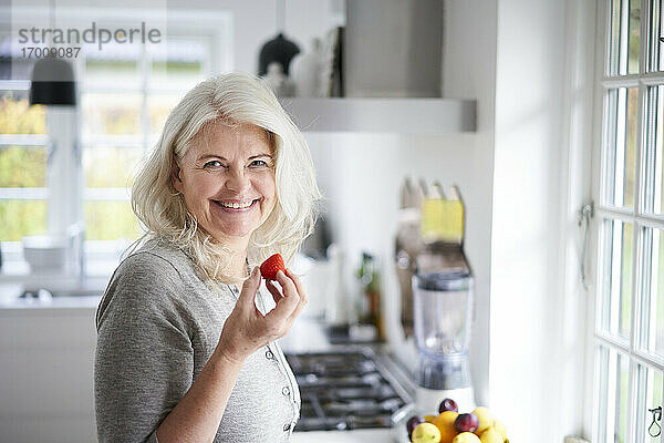 Lächelnde ältere Frau mit langen weißen Haaren  die eine frische Erdbeere in der Hand hält  während sie in der Küche zu Hause steht