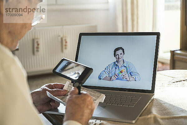 Älterer Mann liest durch ein Vergrößerungsglas  während eine Ärztin per Videoanruf berät