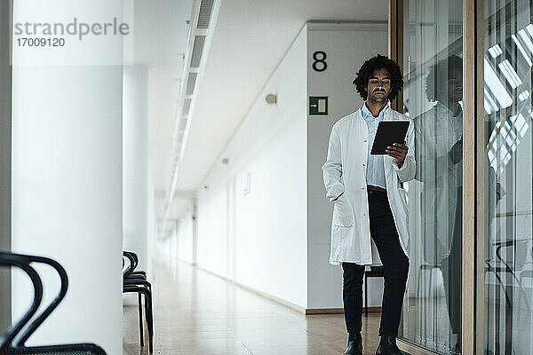 Selbstbewusster junger Arzt  der ein digitales Tablet benutzt  während er sich an eine Glaswand im Krankenhauskorridor lehnt