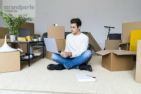 Junger Mann  der einen Laptop benutzt  während er auf dem Boden in einem Raum mit Kisten in einer neuen Wohnung sitzt