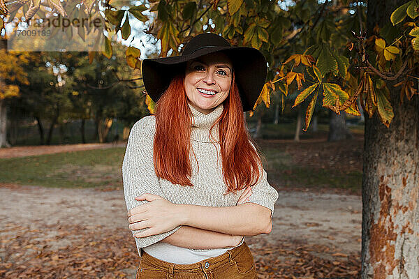Lächelnde schöne Frau mit Hut und verschränkten Armen im Herbst im Park stehend