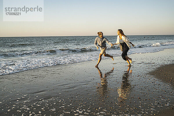 Glückliches Paar hält Hände beim Laufen am Strand gegen den klaren Himmel bei Sonnenuntergang