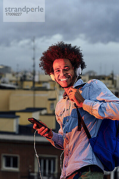 Lächelnder junger Mann mit Rucksack hört Musik über Kopfhörer gegen Gebäude in der Stadt