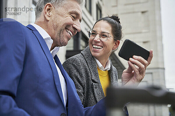 Gut gelaunte Geschäftsfrau  die einen älteren männlichen Kollegen ansieht  der sein Smartphone mit ihr teilt