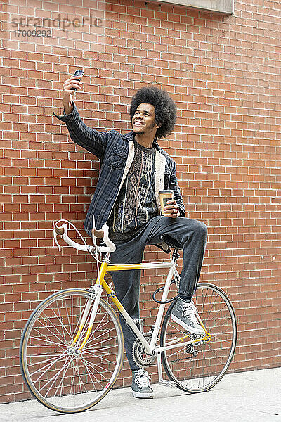 Lächelnder junger Mann mit Kaffee in der Hand  der ein Selfie macht  während er mit dem Fahrrad vor einer Mauer steht