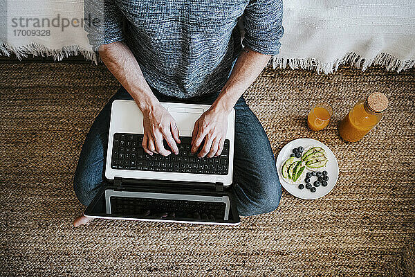 Mann arbeitet am Laptop  während er zu Hause an einem Teller mit Obst und Saft sitzt