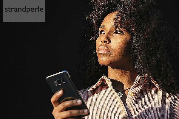 Nachdenkliche afroamerikanische junge Frau  die ihr Handy hält und an einem sonnigen Tag wegschaut